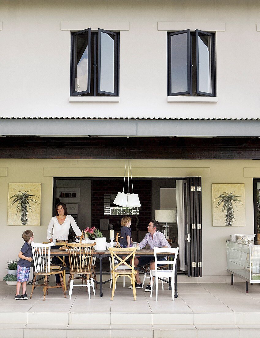 Wohnhaus mit offener Faltschiebetür und überdachter Terrasse, Familie um Tisch mit verschiedenen Stühlen