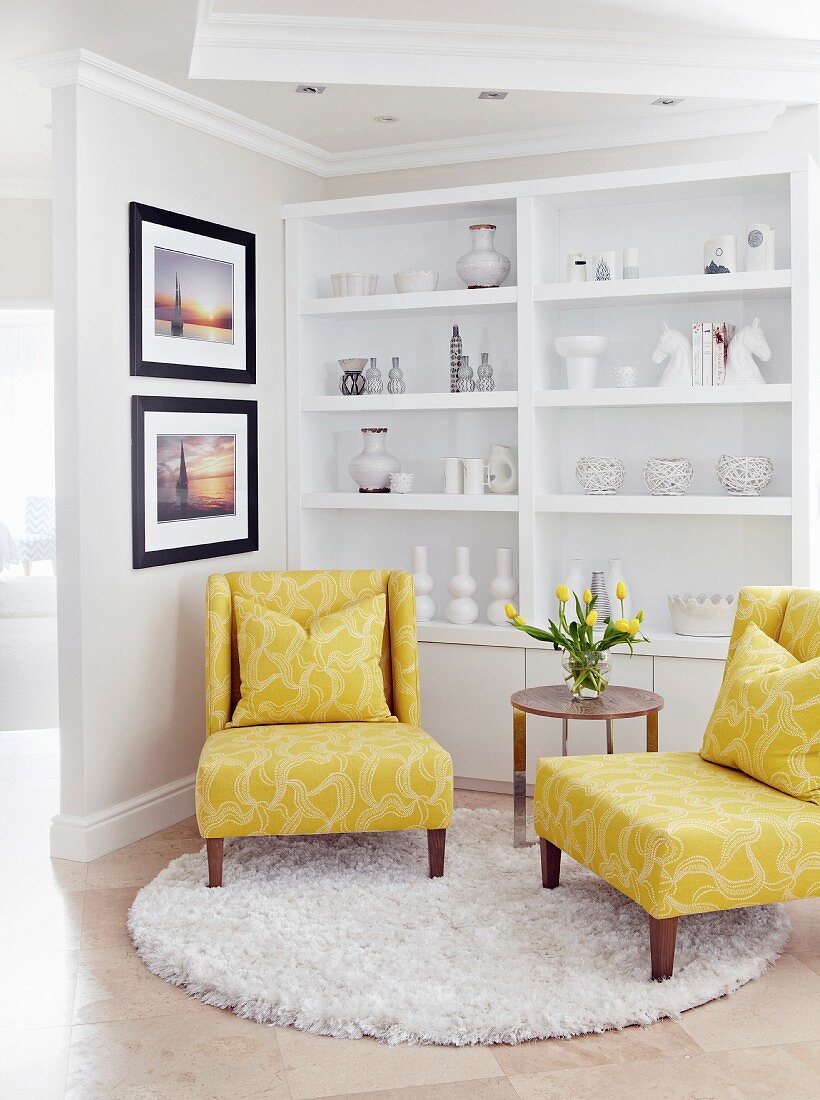 Gelb gepolsterte Sessel mit Beistelltisch auf rundem Teppich, dahinter weisses Regal mit weissen Nippes