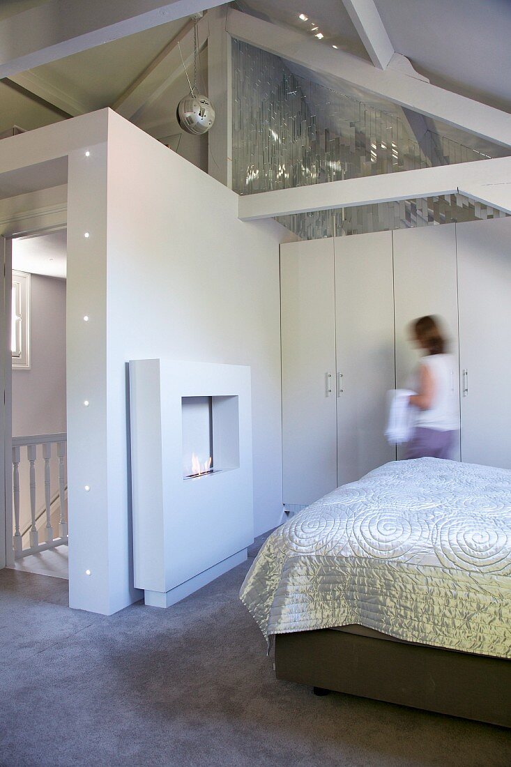 Glossy cover on bed, fake fireplace and woman standing in front of fitted wardrobes in attic bedroom