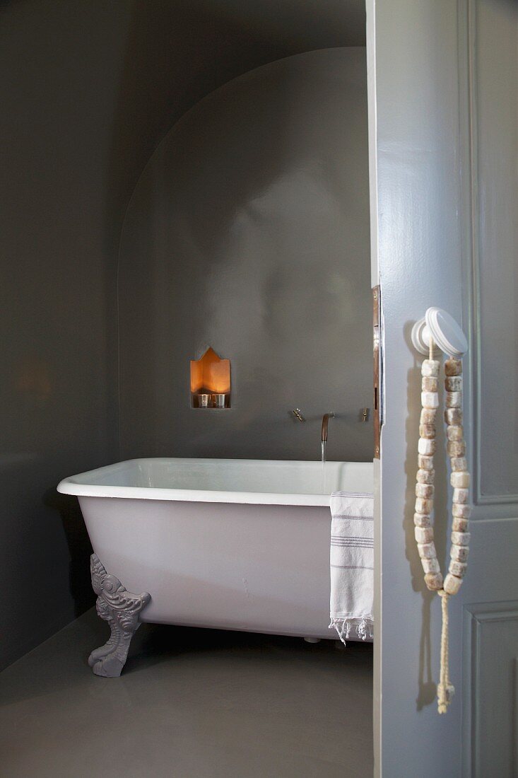String of stone beads hung on handle of open door showing view of free-standing clawfoot bathtub below illuminated niche in dark grey wall