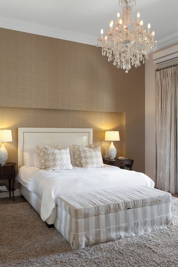 Glamouröses Schlafzimmer in Sandfarben mit gestreifter Bettbank, Kronleuchter und edler Tapete