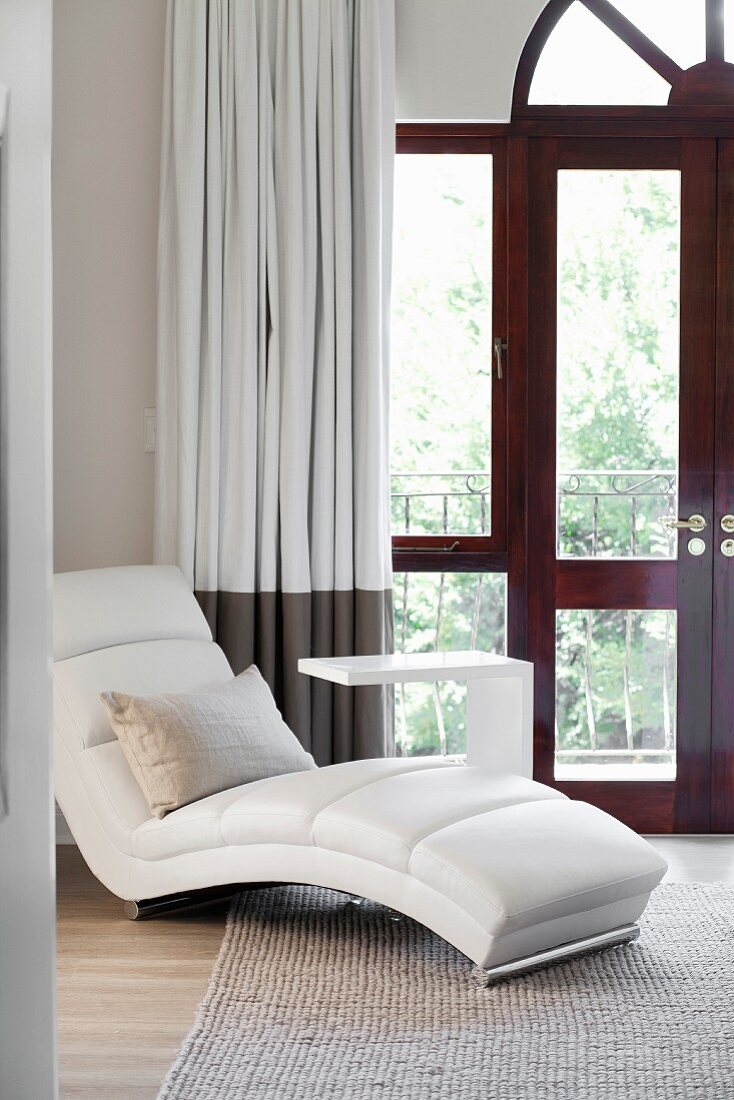 Weißer moderner Polsterliegestuhl mit puristischem Beistelltisch vor Balkontür mit Bogenfenster