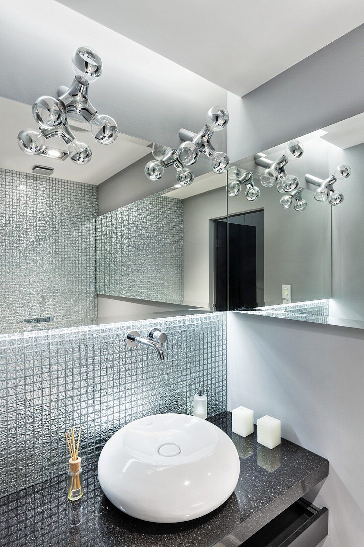Kugelförmiges Waschbecken auf Granitplatte vor Wand mit Mosaikfliesen, oberhalb Designerleuchten