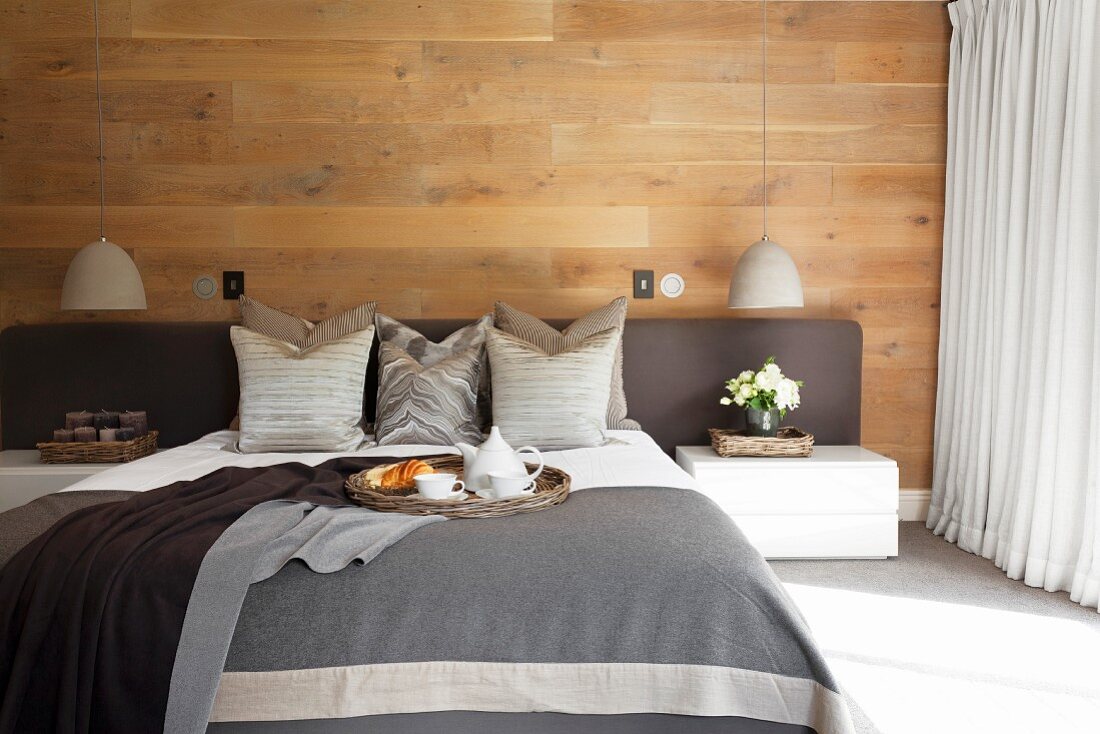 Schlafzimmer mit holzvertäfelter Wand, Doppelbett in Grau mit gepolstertem Kopfteil, Dekokissen und Tagesdecke
