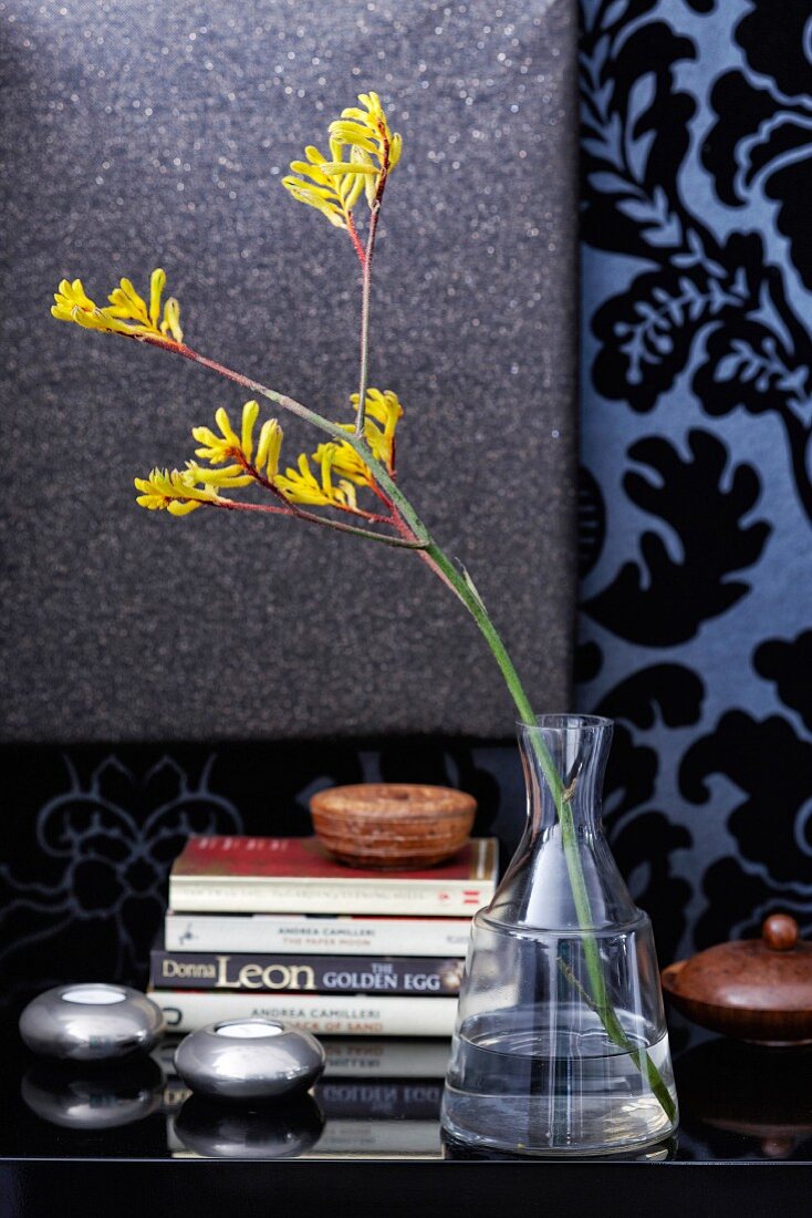 Stacked books, tealight holders and yellow Anigozanthos in glass vase on surface against wall covered in ornate wallpaper