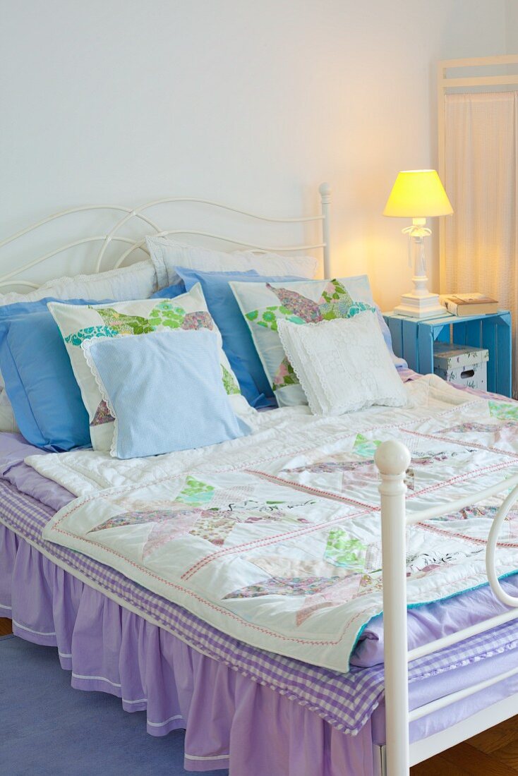Romantisches Doppelbett mit fliederfarbenem Überwurf mit Rüsche; daneben eine pastellblau lackierte Obstkiste als Nachttisch