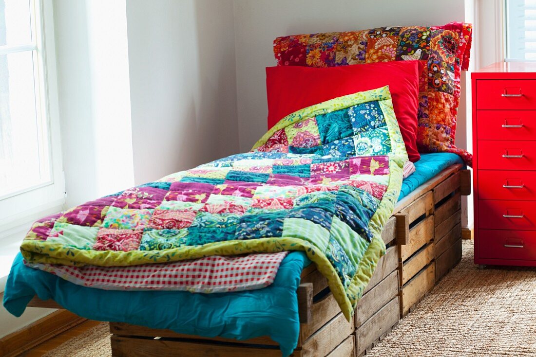 Rustikales, aus Holzkisten gebautes Bett mit bunten Patchworkdecken; daneben ein leuchtend roter Metallcontainer