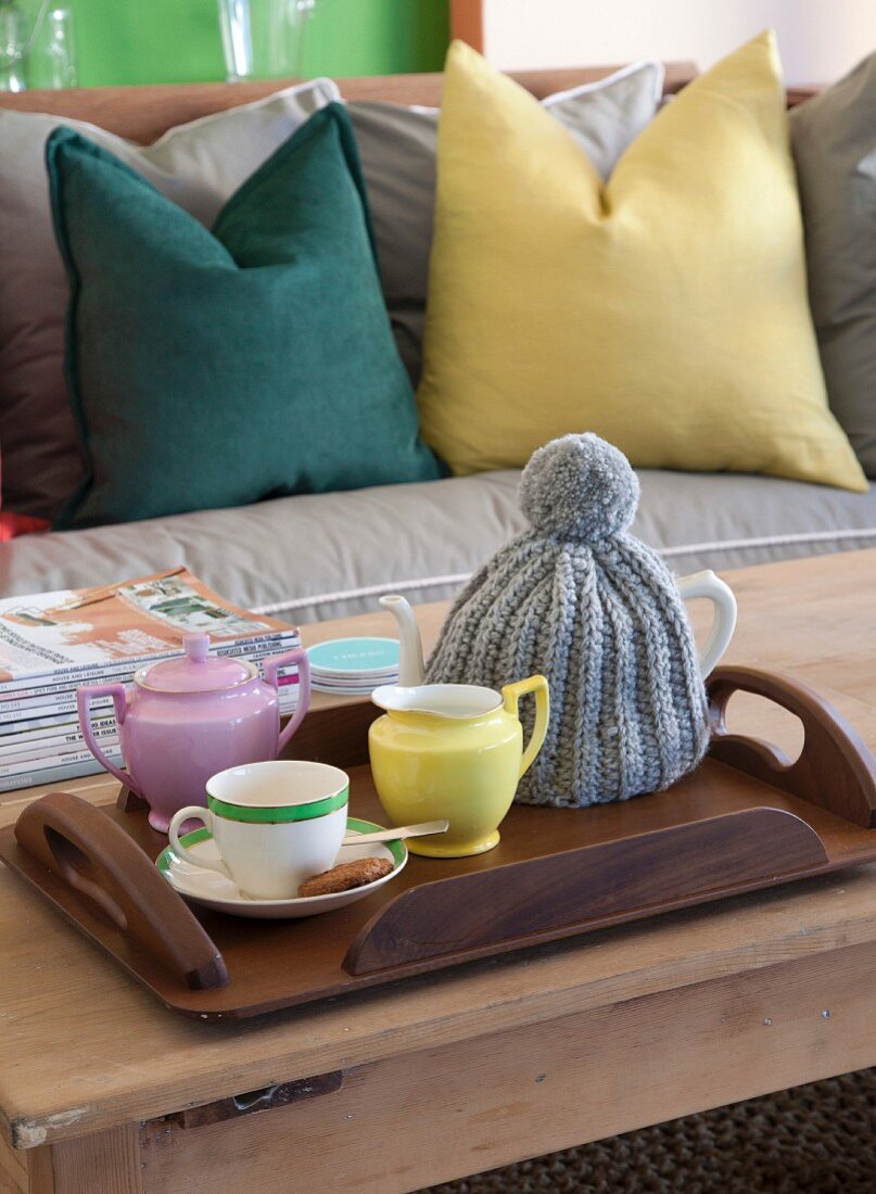 Teekanne mit gestricktem Wärmer und Tasse auf Tablett, im Hintergrund farbige Kissen auf Sofa