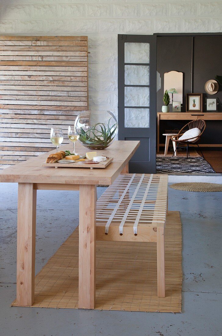Narrow wooden table and bench with woven seat on terrace in front of open double doors with view of retro armchair