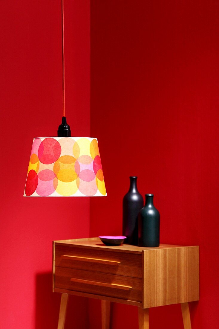 Schlichter Lampenschirm mit bunten Kreisen aufgepeppt, vor Retro Kommode mit Keramikflaschen an roter Wand