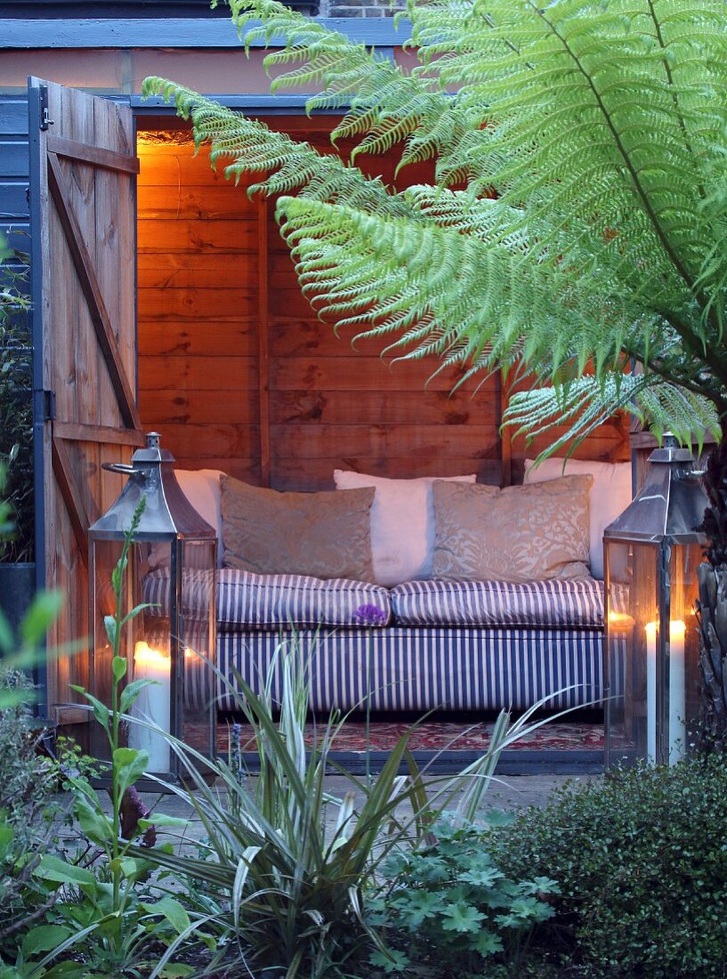 Illuminated wooden shed with sofa, two lanterns and tree fern in garden