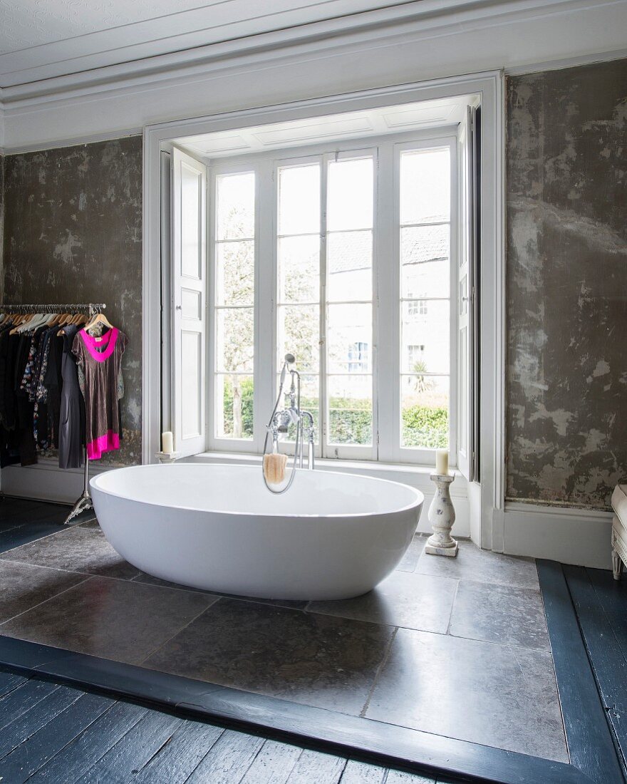 Oval, free-standing designer bathtub on stone tiles in front of bedroom window