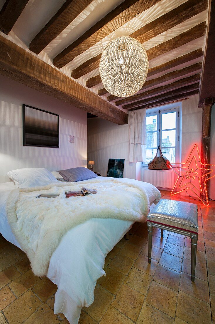 Weisses Fell auf Doppelbett unter Kugelleuchte, im Hintergrund sternförmige Leuchte in Rot in rustikal elegantem Schlafzimmer mit Holzbalkendecke