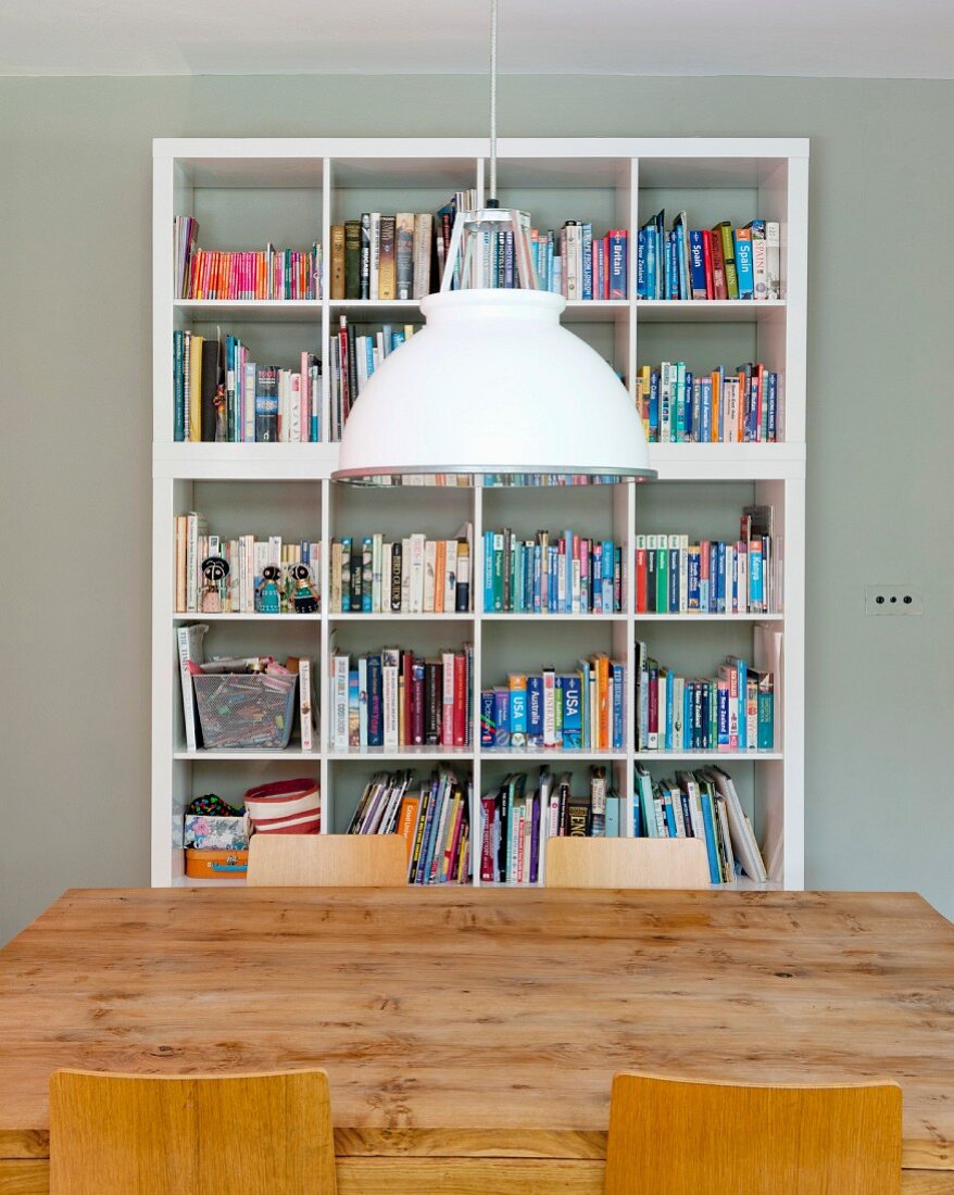 Pendant lamp with white-painted metal lampshade above wooden dining table in front of white bookcase against wall painted pale grey