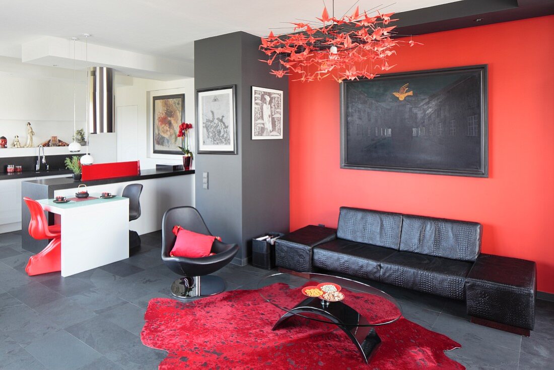 Black leather couch below artwork on red wall, coffee table on red rug and tiled floor and kitchen area in background