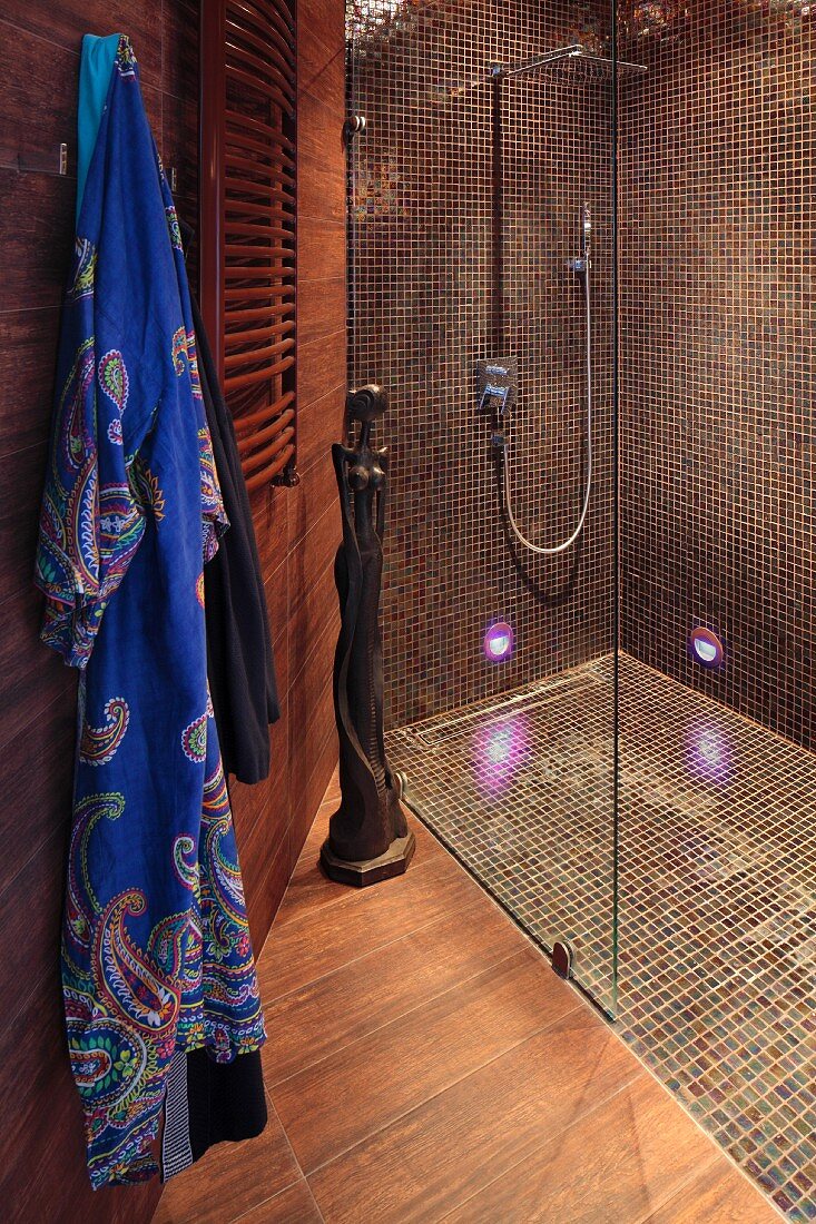 Duschbereich mit Glastrennscheibe, braunen Mosaikfliesen an Wand und Boden, seitlich aufgehängter Morgenmantel im Designerbad