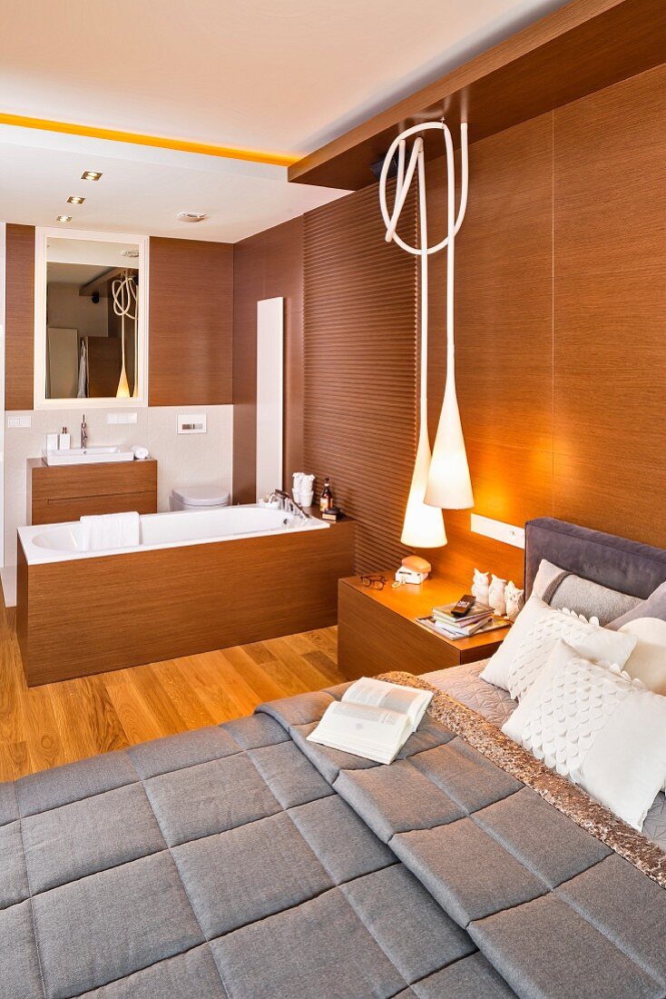 Elegant bed against wood-clad wall and designer pendant lamps in bedroom with ensuite bathroom
