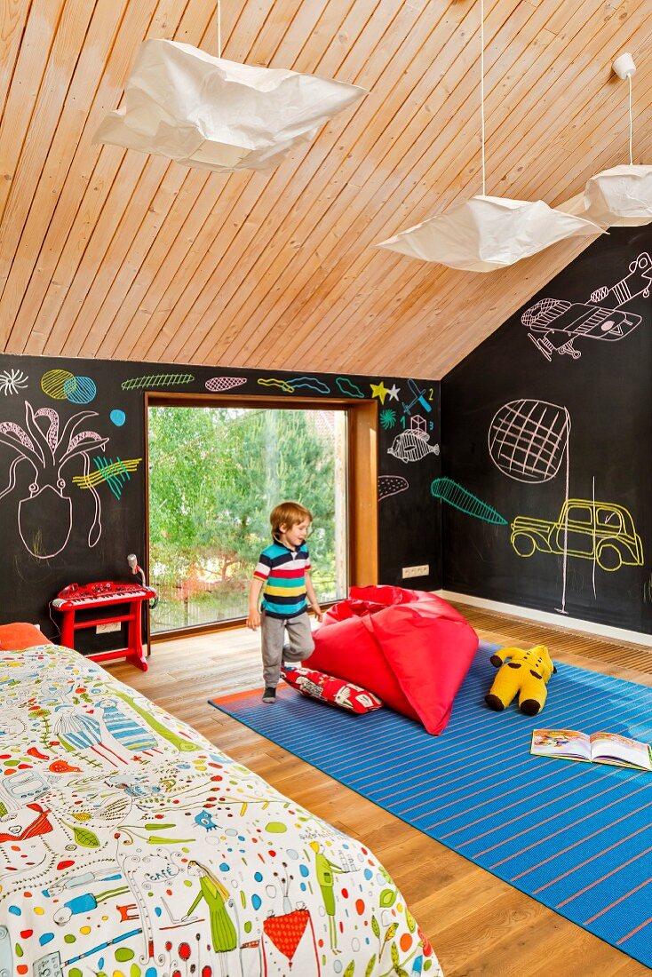 Kleiner Junge neben rotem Sitzsack auf blauem Teppich, an Wand Tafellack und Kreidezeichnungen, im Kinderzimmer unter dem Dach, holzverkleidete Decke