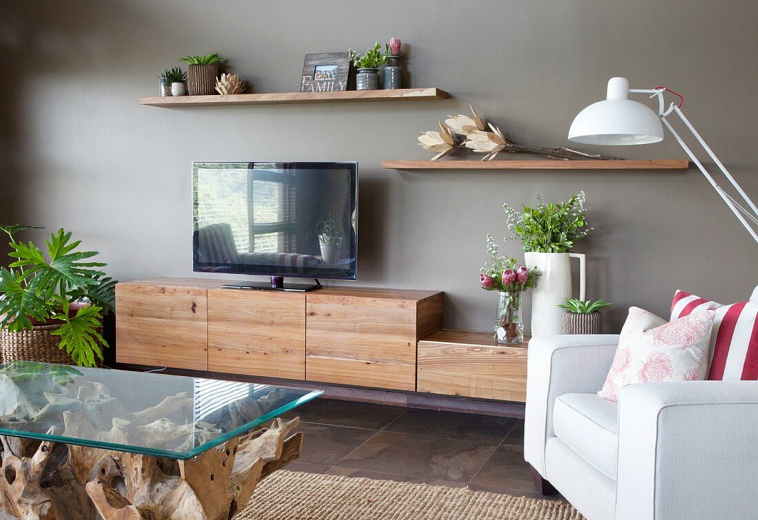 Couchtisch mit Glasplatte und heller Sessel, gegenüber Lowboard mit rustikalen Schrankmodulen aus Holz an grauer Wand
