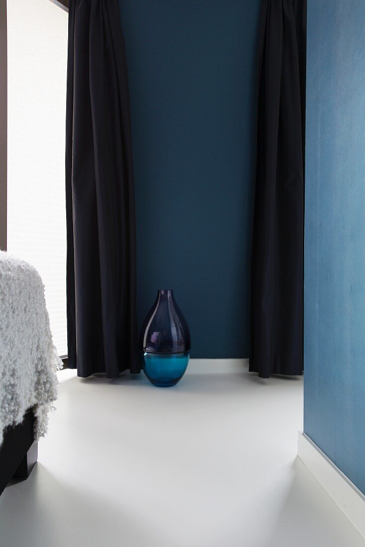 Bodenvase aus blauem Glas auf weißem Epoxidharzboden, zwischen dunkelblauen, bodenlangen Vorhängen