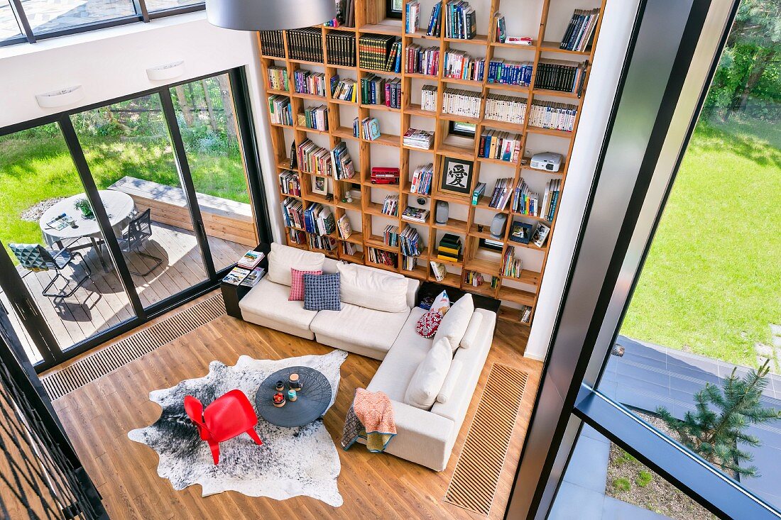 Blick von oben auf Wohnzimmer, helle Sofagarnitur und roter Klassikerstuhl auf Tierfell, an Wand raumhohes Bücherregal aus Holz