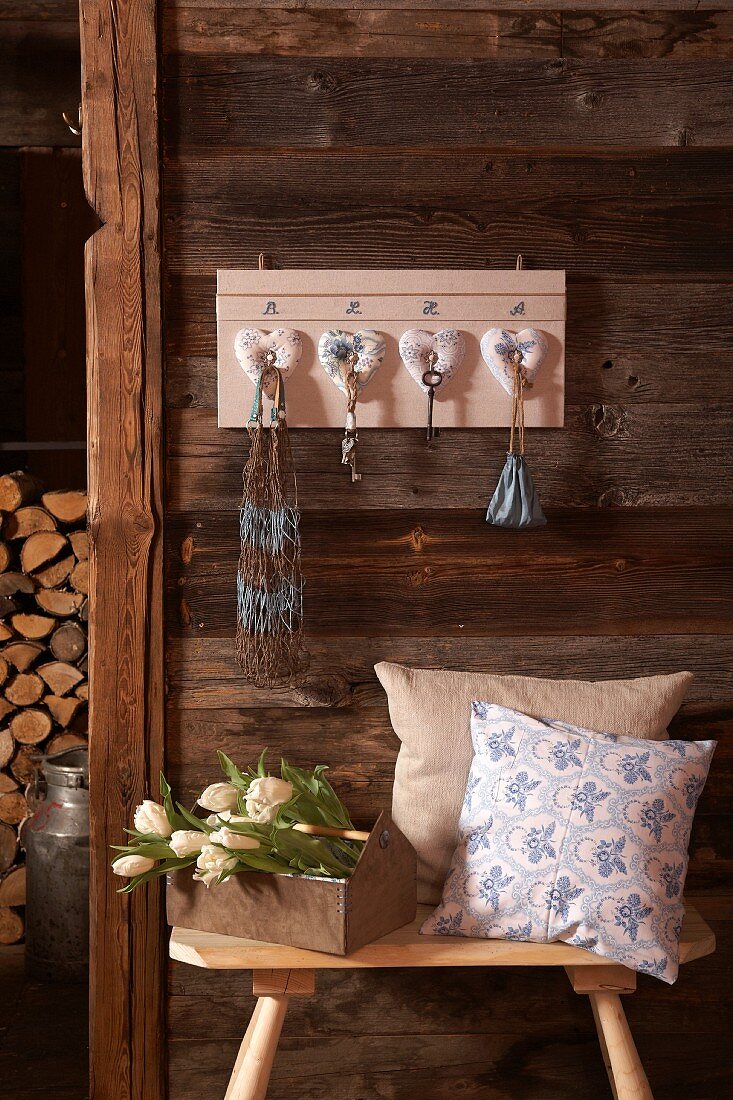 DIY-Schlüsselbrett mit Herzdeko an Holzwand, davor Schemel mit Kissen und Blumenstrauss