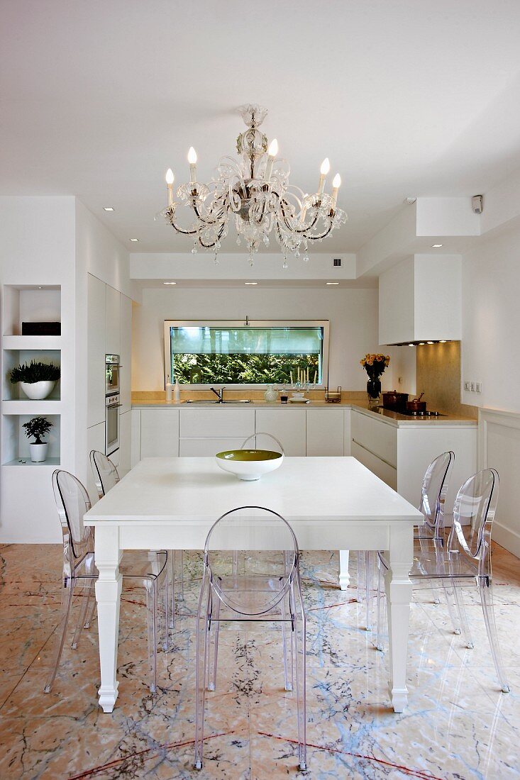 Ghost chairs around white, country-house-style dining table below chandelier in open-plan kitchen