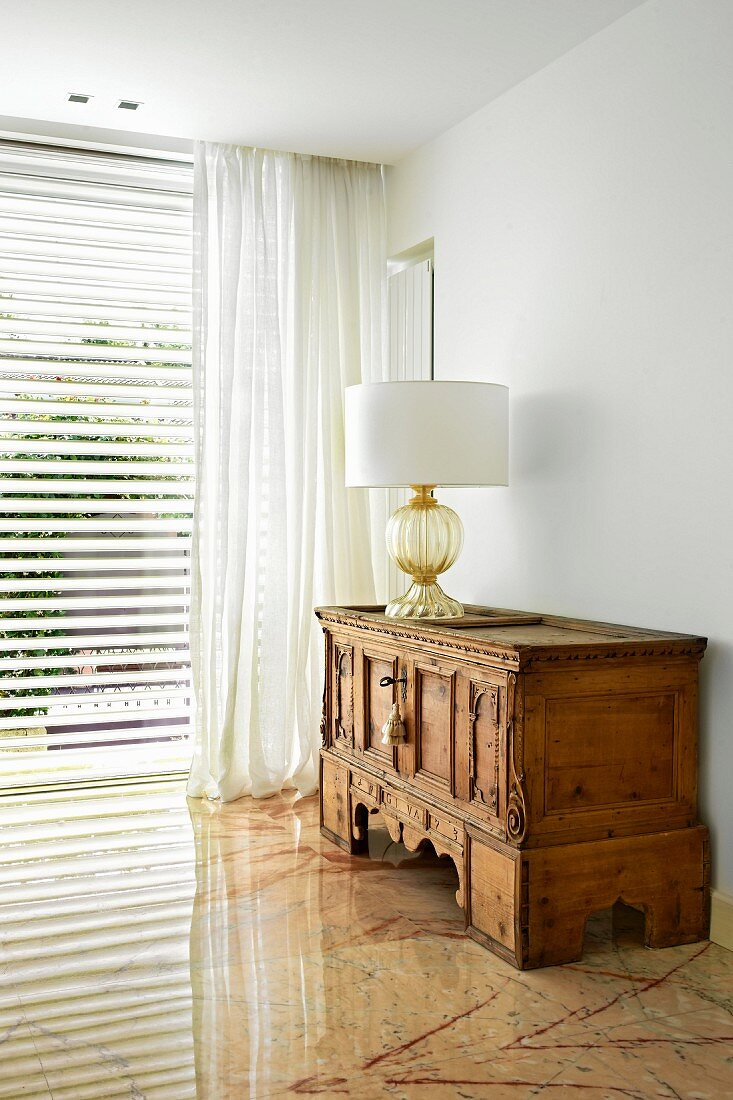 Table lamp with white lampshade on antique, carved wooden cabinet in corner of living room with marble floor