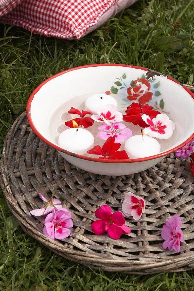 Emailleschüssel mit Schwimmkerzen und Geranienblüten auf Rattanuntersetzer
