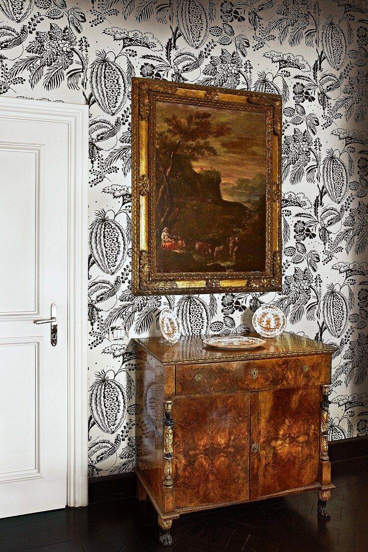 Antike Kommode mit Schnitzereien und gerahmtes Gemälde vor tapezierter Wand mit floralem Mustern
