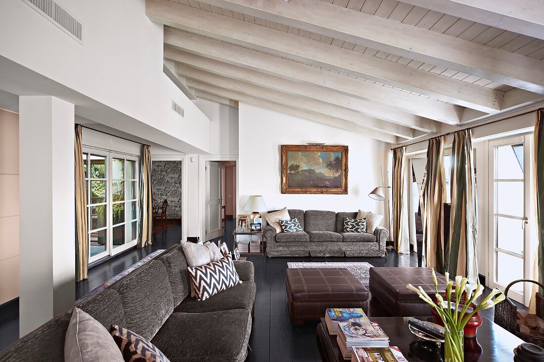Grey couch and ottomans in seating area of open-plan interior with white-painted wooden ceiling