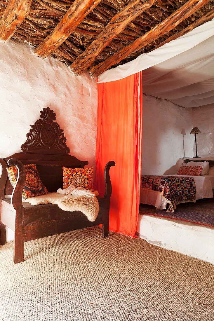 Antike Sitzbank mit Schnitzerei auf Sisalboden, orangefarbener Vorhang vor Schlafbereich auf Podest