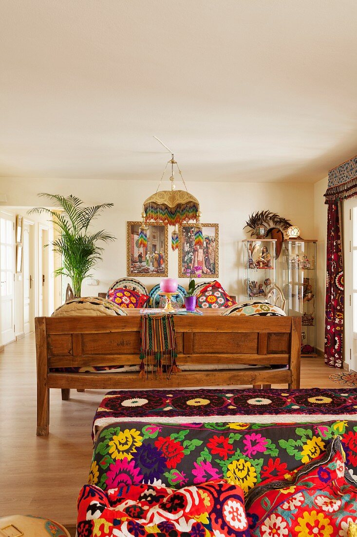 Sofa with various brightly patterned textiles and wooden bench in comfortable, ethnic-style living room
