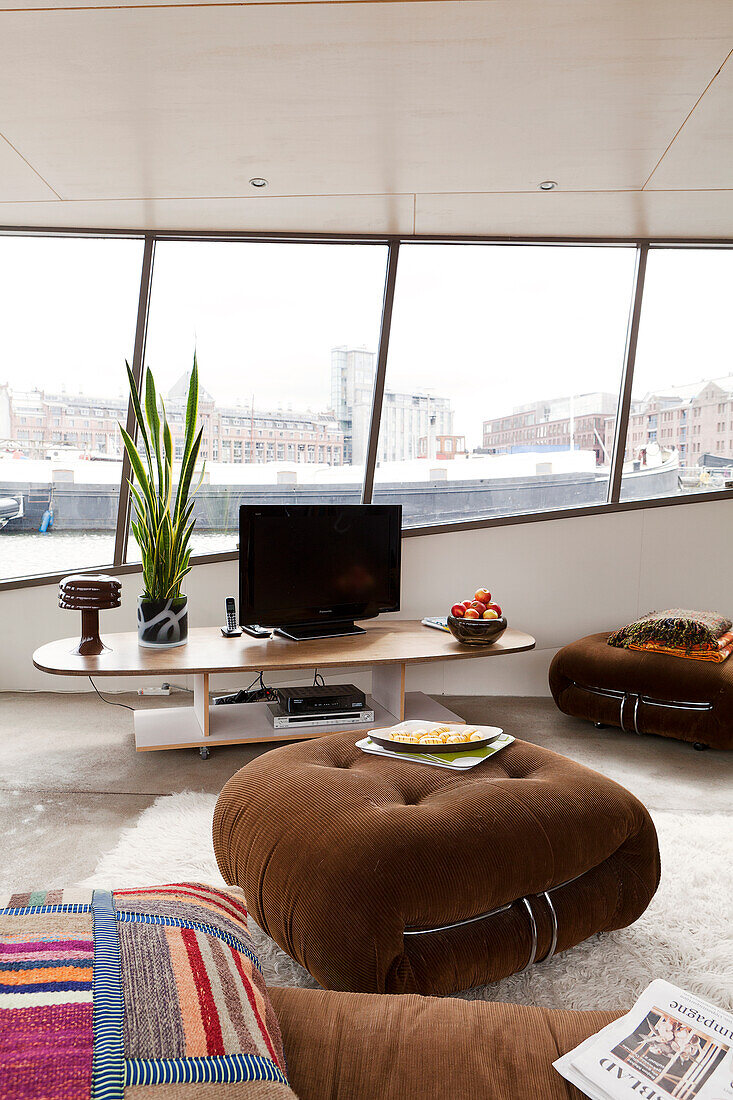 Brown ottoman in front of flatscreen TV on low table with view of harbour though strip of windows in background