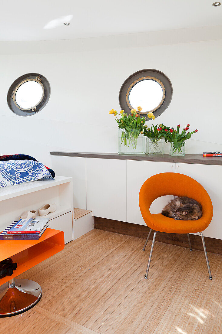 Cat sitting on orange retro armchair in bedroom of houseboat with custom fitted furniture and porthole windows