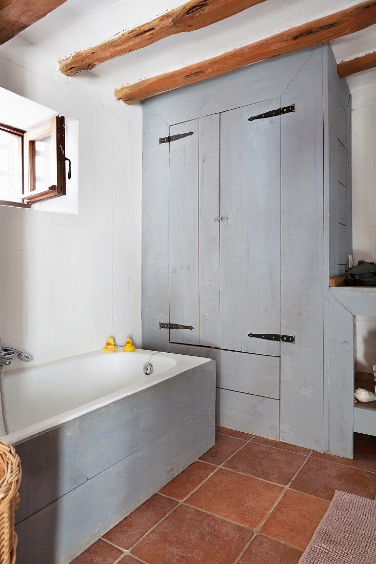 Bathtub with pale grey wooden side and matching fitted cupboard in rustic bathroom with terracotta floor tiles