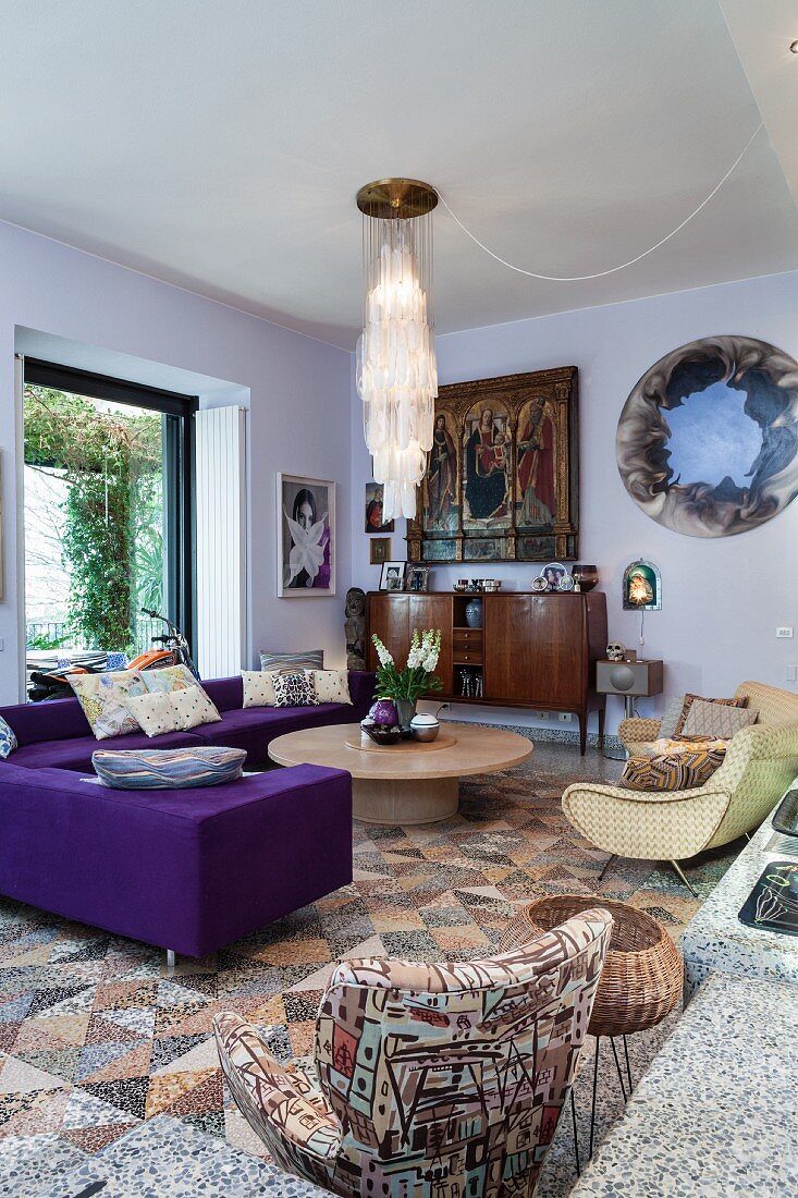 Wohnzimmer mit violettem Sofa und Retro Sessel um rundem Couchtisch, unter moderner Pendelleuchte
