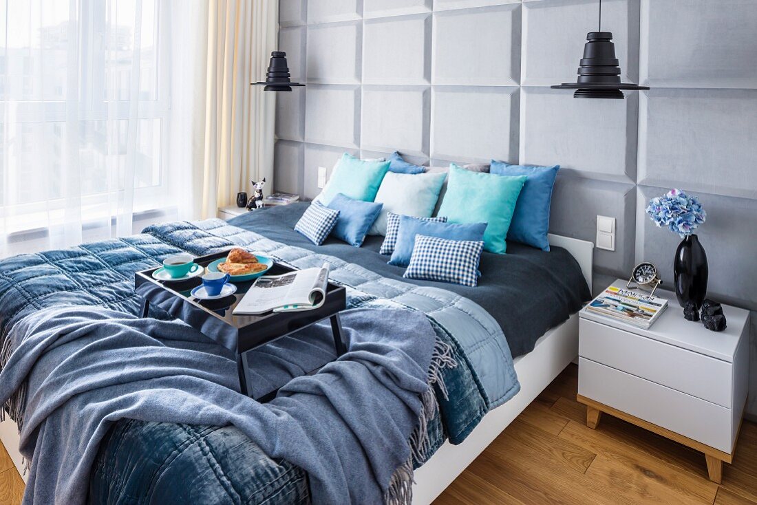 Frühstückstablett auf Doppelbett mit Kissen und Decken in Blautönen; hellgraue Rückwand mit Kassettenstruktur