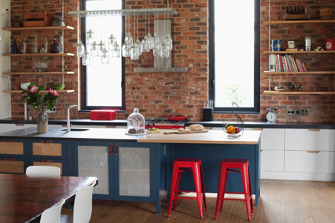 Rote Metallhocker an Kücheninsel mit viel Stauraum; Küchenzeile vor Backsteinwand