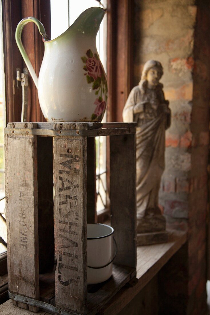 Alter Waschkrug auf Holzkiste, im Hintergrund religiöse Figur, auf Fensterbank