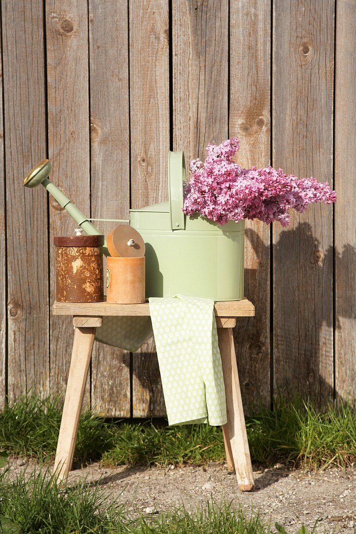 Pastellgrüne Giesskanne mit Fliederstrauss und Aufbewahrungsbehälter auf Holzschemel, im Freien vor Holzwand