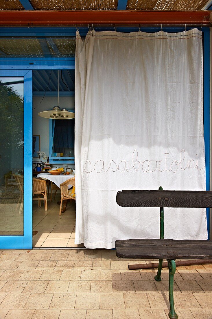 Weisser Vorhang vor geöffneter, hellblauer Glasschiebetür auf südländischer Terrasse
