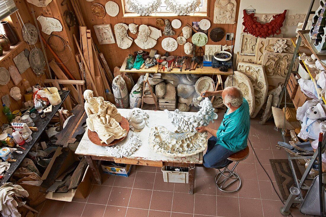 View down onto sculptor at workbench making sculptures and masks in workshop