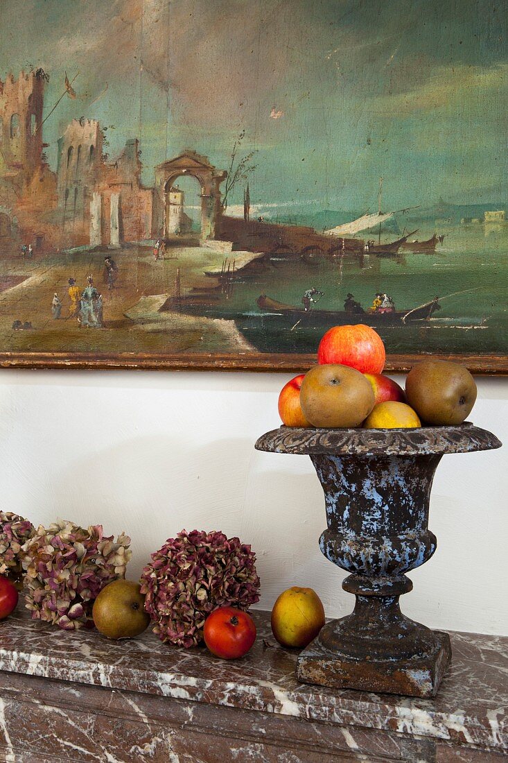 Hortensienblüten, Äpfel und antikes Pokalgefäss als Deko auf Marmor-Kaminsims unter Wandgemälde