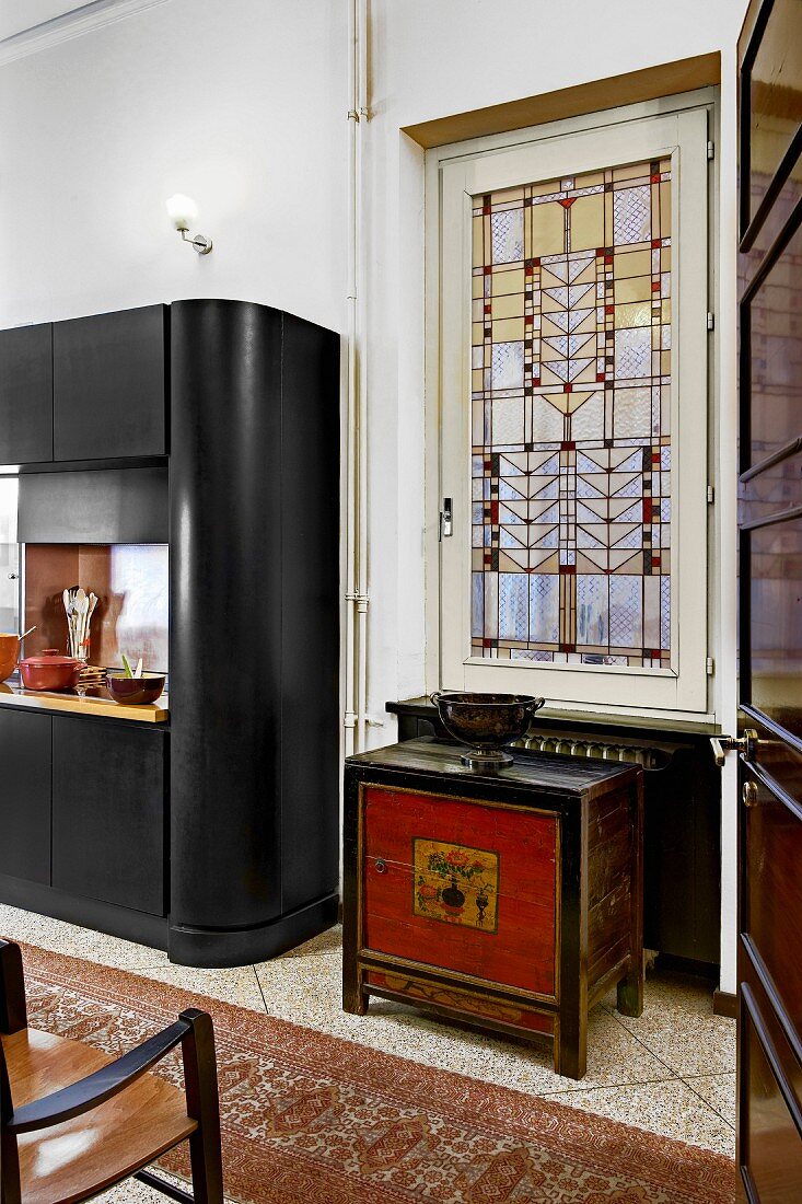Painted, half-height cabinet below window with art-nouveau stained glass; detail of modern, black kitchen cupboard to one side