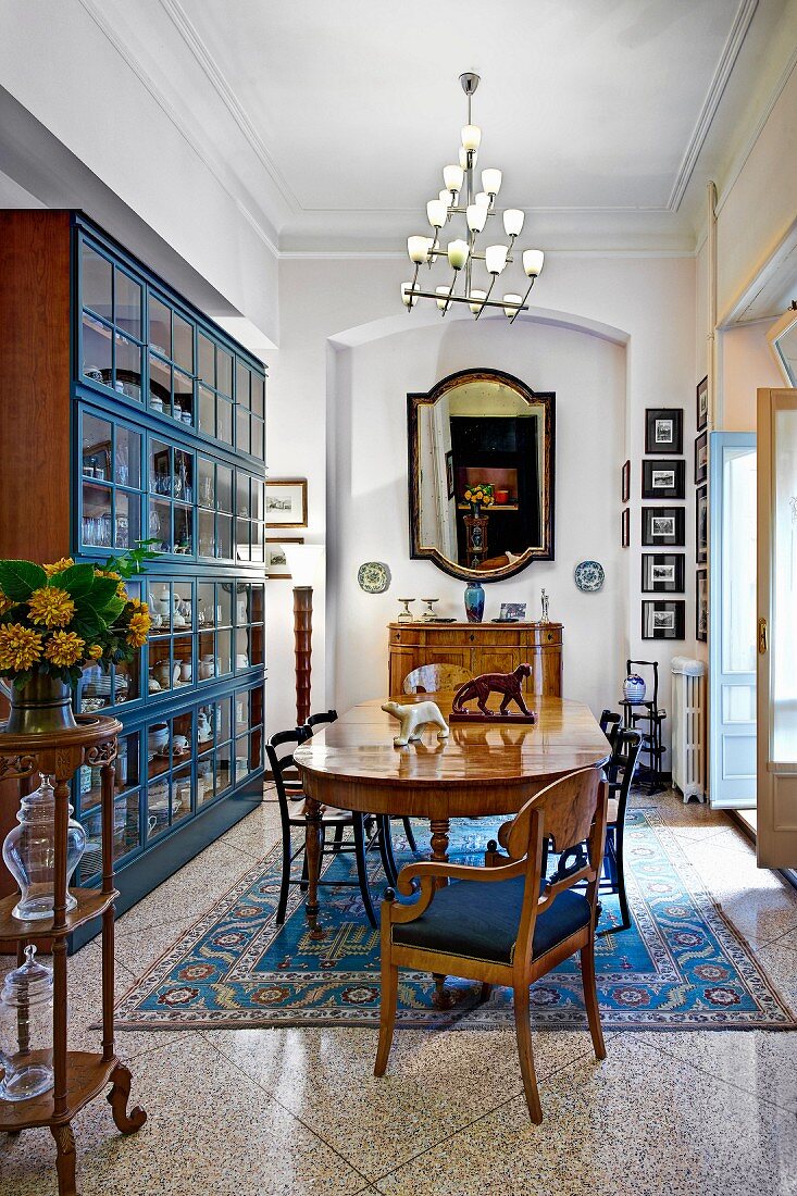 Antike Stühle und Tisch auf gemustertem Teppich, gegenüber Vitrinenschrank mit blau lackiertem Rahmen, in traditionellem Ambiente, Pendelleuchte mit kleinen Glasschirmen
