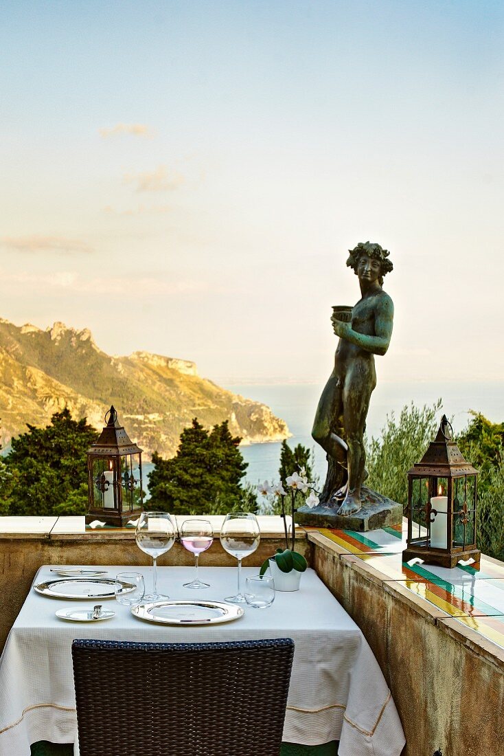 Skulptur auf Terrassen-Brüstungsmauer, davor gedeckter Tisch für Zwei, im Hintergrund Meeresküste