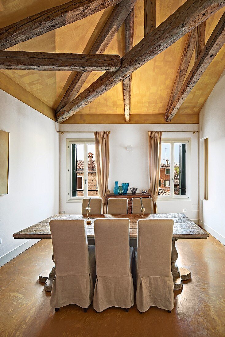 Dining area with rustic wooden table and pale, loose-covered chairs below exposed wooden roof beams
