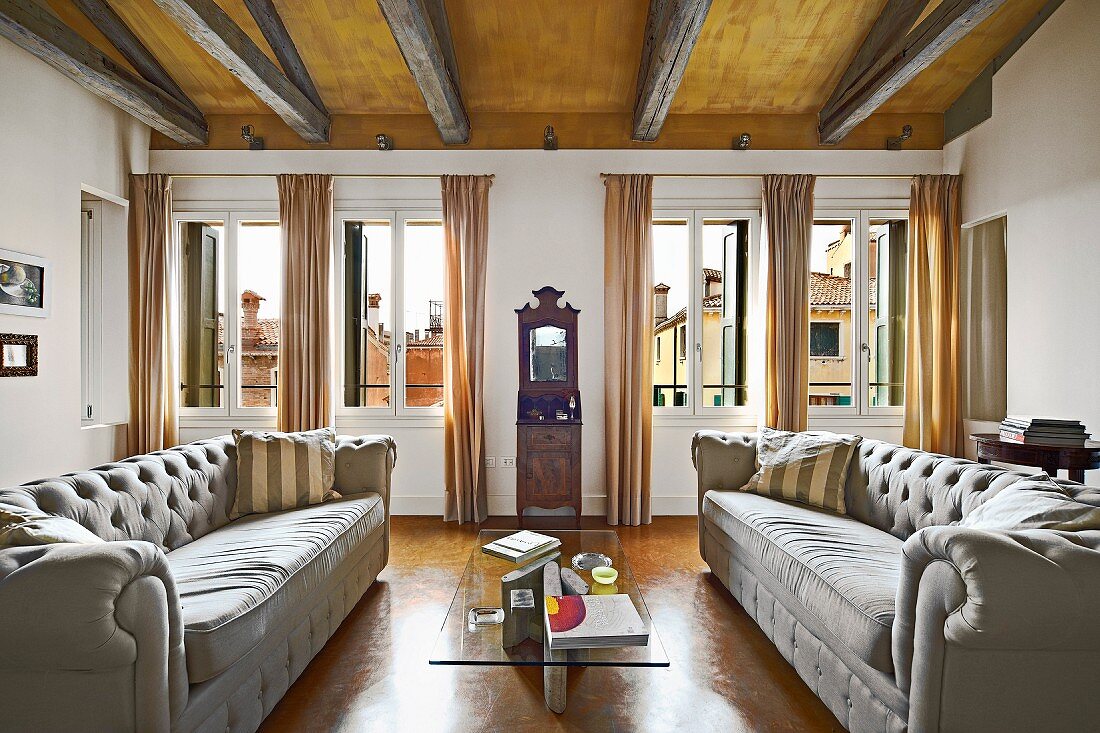 Helle kapitonierte Couchgarnitur vor Fensterfront mit bodenlangen Vorhängen in Wohnzimmer mit italienischem Flair