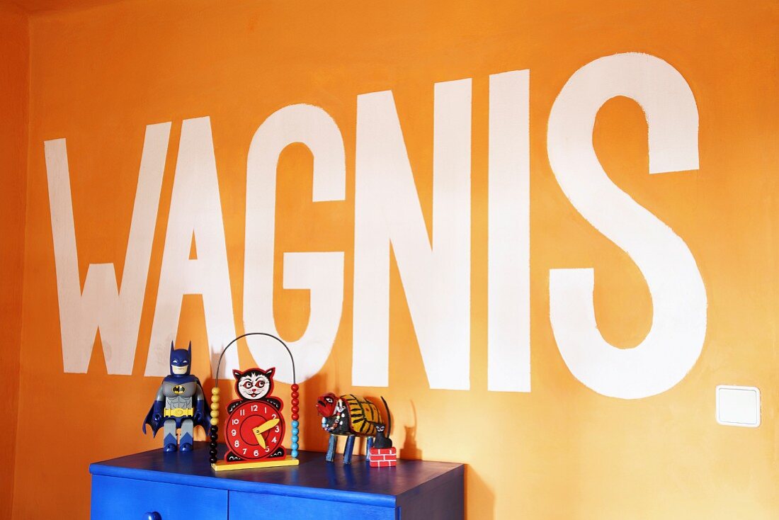 Botschaft auf orangefarbener Wand, davor teilweise sichtbarer blauer Schrank mit Vintage Spielsachen